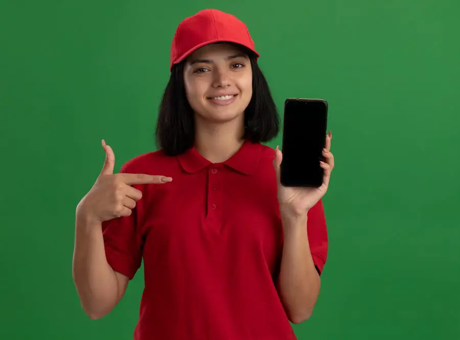 young delivery girl red uniform cap showing smartphone pointing with index finger it smiling confident standing green wall