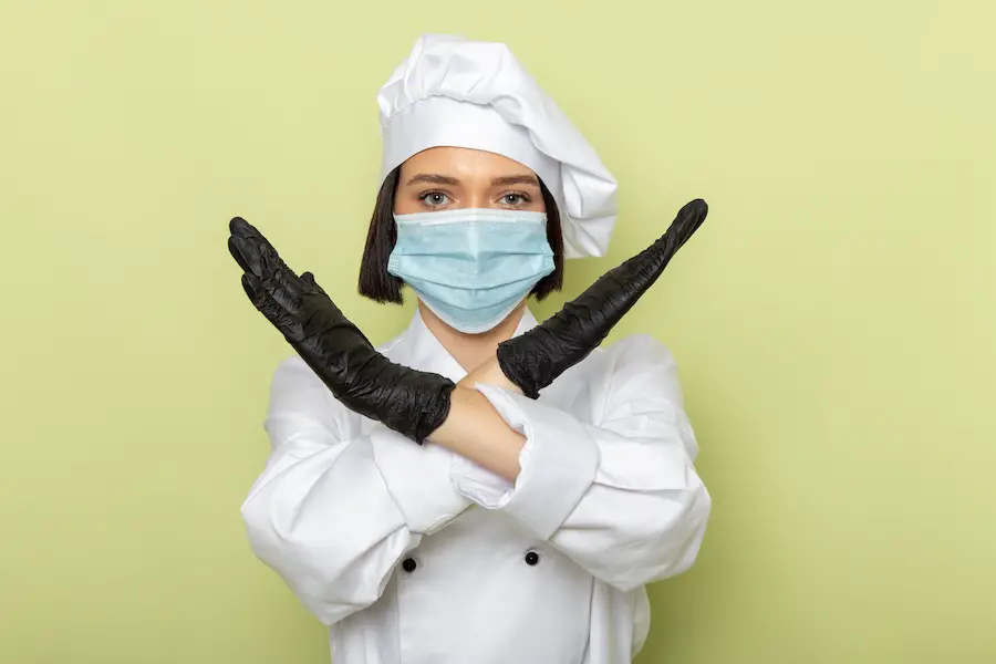 a front view young female cook in white cook suit and cap wearing gloves and sterile mask with ban pose on the green background lady work food color